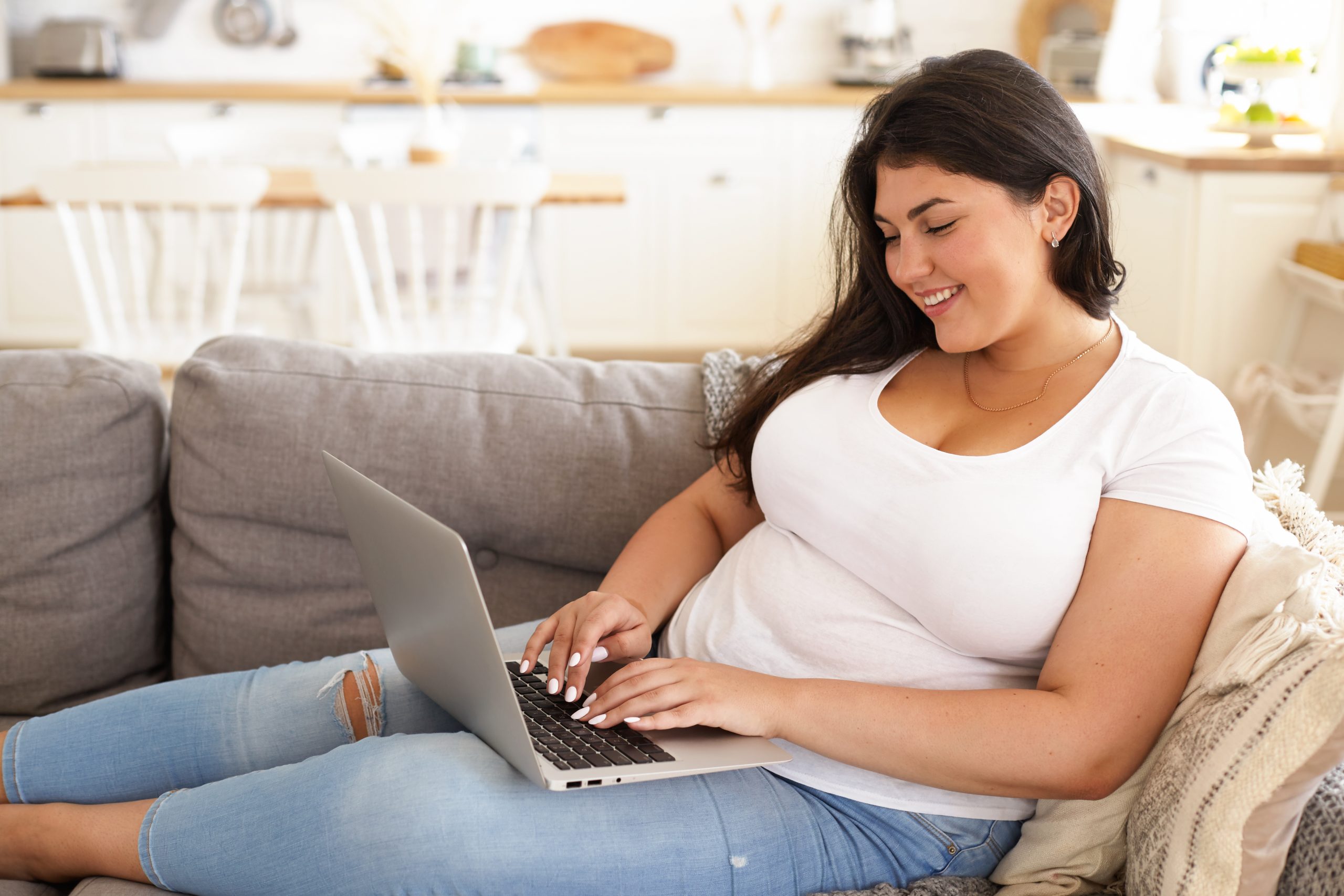 Side view of fashionable young overweight plump female freelancer sitting on sofa with portable computer on her lap, typing fast, working remotely from home. Brunette plus size girl using laptop
